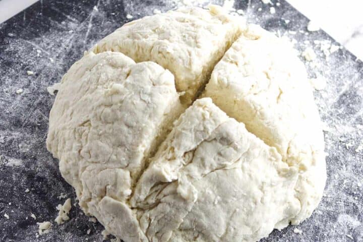 A ball of raw biscuit dough on a floured surface with a large cross-shaped cut in the center, just like in a traditional Irish soda bread recipe, ready to be divided or baked.