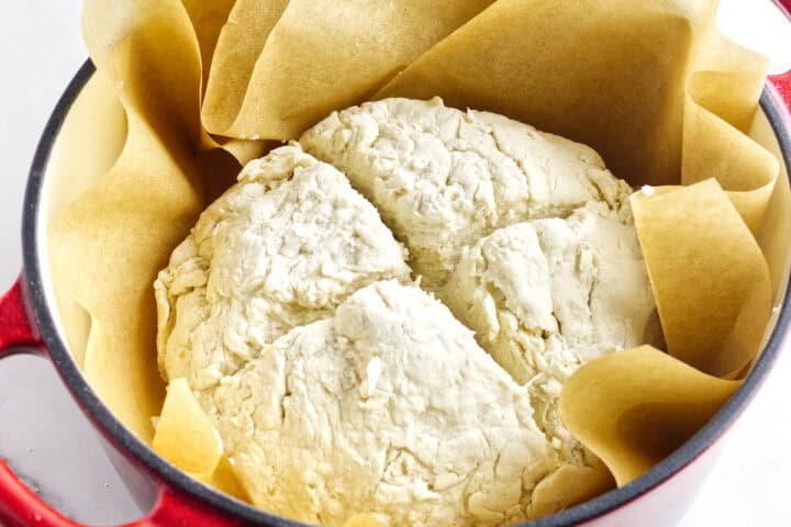 A round loaf of unbaked bread dough sits on parchment paper inside a red pot, ready to be baked. The dough appears slightly cracked, hinting at a rustic texture perfect for a traditional Irish soda bread recipe.