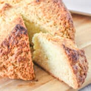 A close-up of a round, golden-brown loaf made from a traditional Irish soda bread recipe sits on a wooden surface. One thick slice is cut and slightly pulled away, revealing the soft, airy interior beneath its rustic crust.