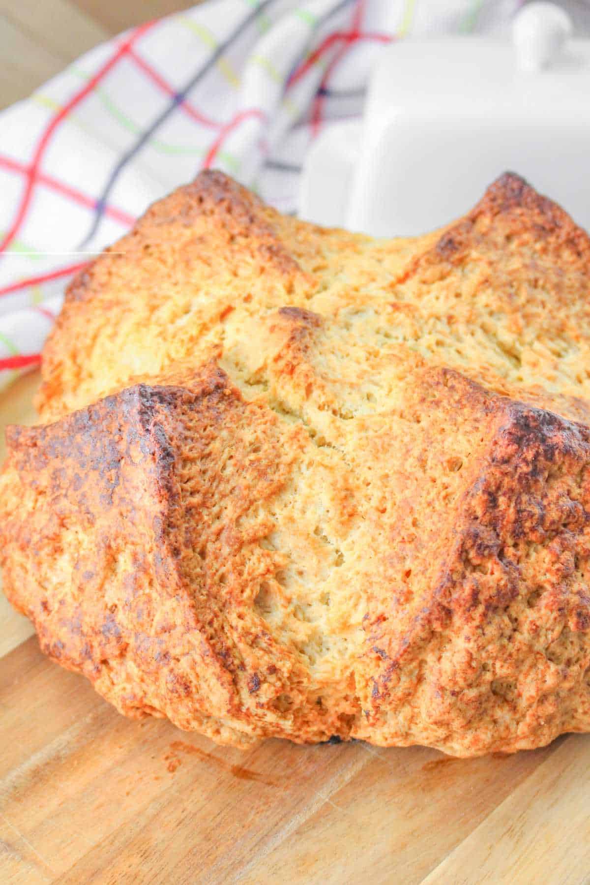 A freshly baked round loaf made from a traditional Irish soda bread recipe rests on a wooden surface, its golden-brown crust shining with warmth, while a colorful kitchen towel sits in the background.