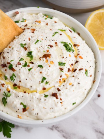 A bowl of creamy whipped feta dip garnished with chopped parsley, red pepper flakes, black pepper, and a drizzle of olive oil, with a pita chip dipping in. A lemon slice and parsley sit nearby on the marble surface.