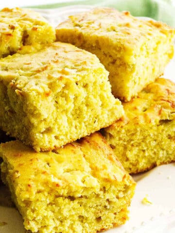 Golden, fluffy pieces of cornbread are stacked on a white plate, showcasing their moist and crumbly texture with a slightly crisp, browned top.