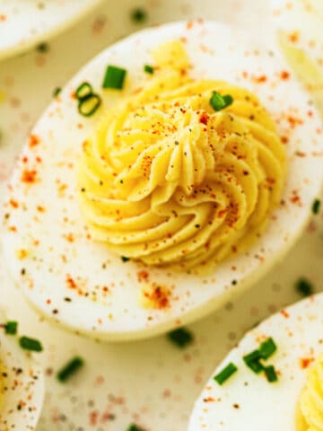 Close-up of deviled eggs topped with a creamy, piped yolk filling, garnished with chopped chives and a sprinkle of paprika and black pepper on a white surface-perfect for deviled egg lovers.