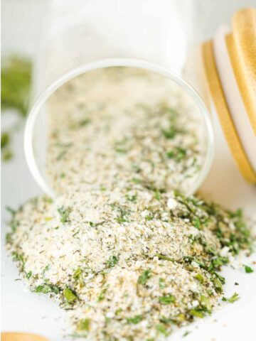 A glass jar tipped on its side spills out a mixture of dried herbs and breadcrumbs, resembling a homemade dry Ranch seasoning mix recipe, onto a white surface, with chopped green herbs visible in the blend.
