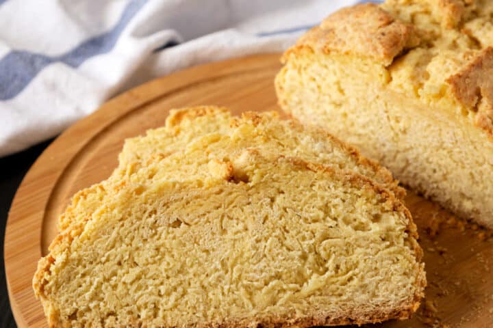 Close-up of two slices of golden brown, crusty gluten free Irish soda bread on a wooden cutting board, with a white and blue cloth in the background.