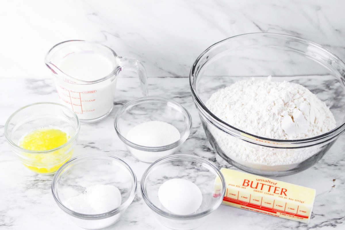 A collection of gluten free baking ingredients on a marble surface: a bowl of flour, butter, milk, sugar, an egg, melted butter, and baking powder—perfect for making Irish Soda Bread.