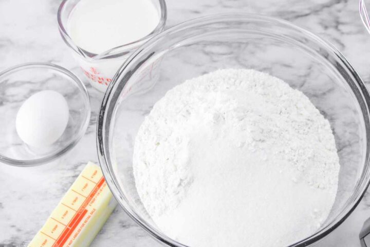 A glass bowl of flour and sugar, a stick of butter, a whole egg in a small bowl, and a measuring cup of milk are arranged on a marble countertop for making gluten free Irish soda bread.