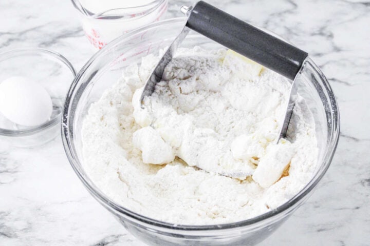 A glass bowl with flour and chunks of butter being mixed with a pastry cutter for gluten free Irish soda bread. Nearby, there is a clear bowl with an egg and a measuring cup with milk, all placed on a marble countertop.