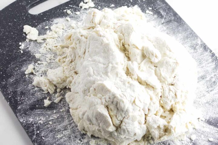 A mound of rough, partially mixed gluten free dough sits on a floured black cutting board, with small bits of flour and dough scattered around it—perfect for crafting homemade gluten free Irish soda bread.