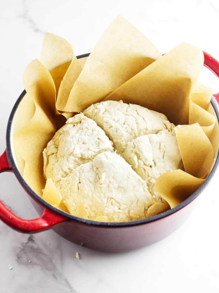 A round loaf of rustic, partially cracked Gluten Free Bread sits on brown parchment paper inside a red Dutch oven on a white surface.