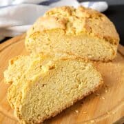 A round loaf of golden-brown gluten free Irish soda bread with a crunchy crust is sliced on a wooden cutting board, showing its dense, crumbly interior. A striped cloth is in the background.