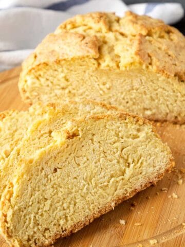 A round loaf of golden-brown gluten free Irish soda bread with a crunchy crust is sliced on a wooden cutting board, showing its dense, crumbly interior. A striped cloth is in the background.