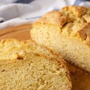 A close-up of two slices of golden, crusty gluten free Irish soda bread on a wooden cutting board, with a white and blue striped cloth in the background.