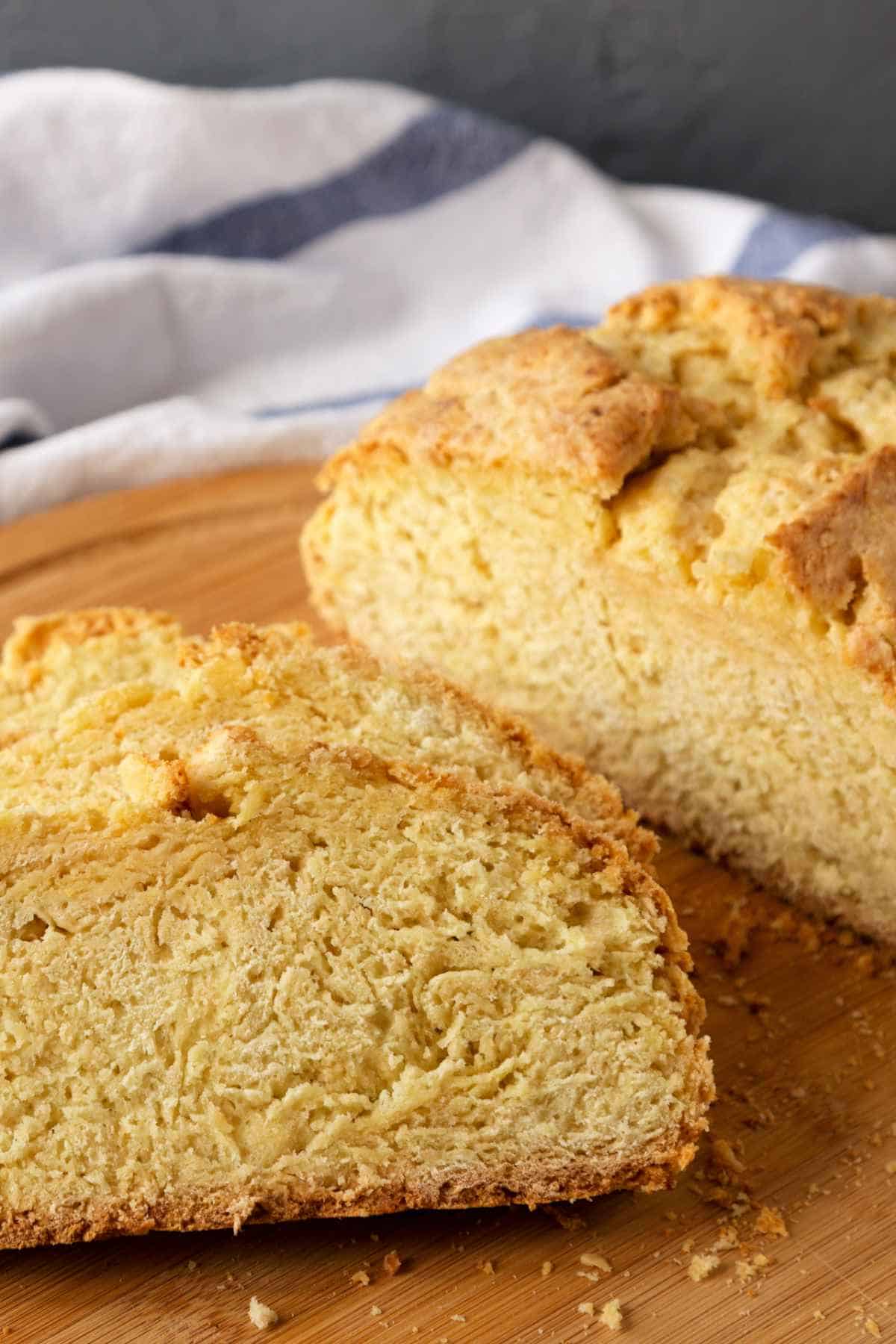 A close-up of two slices of golden, crusty gluten free Irish soda bread on a wooden cutting board, with a white and blue striped cloth in the background.