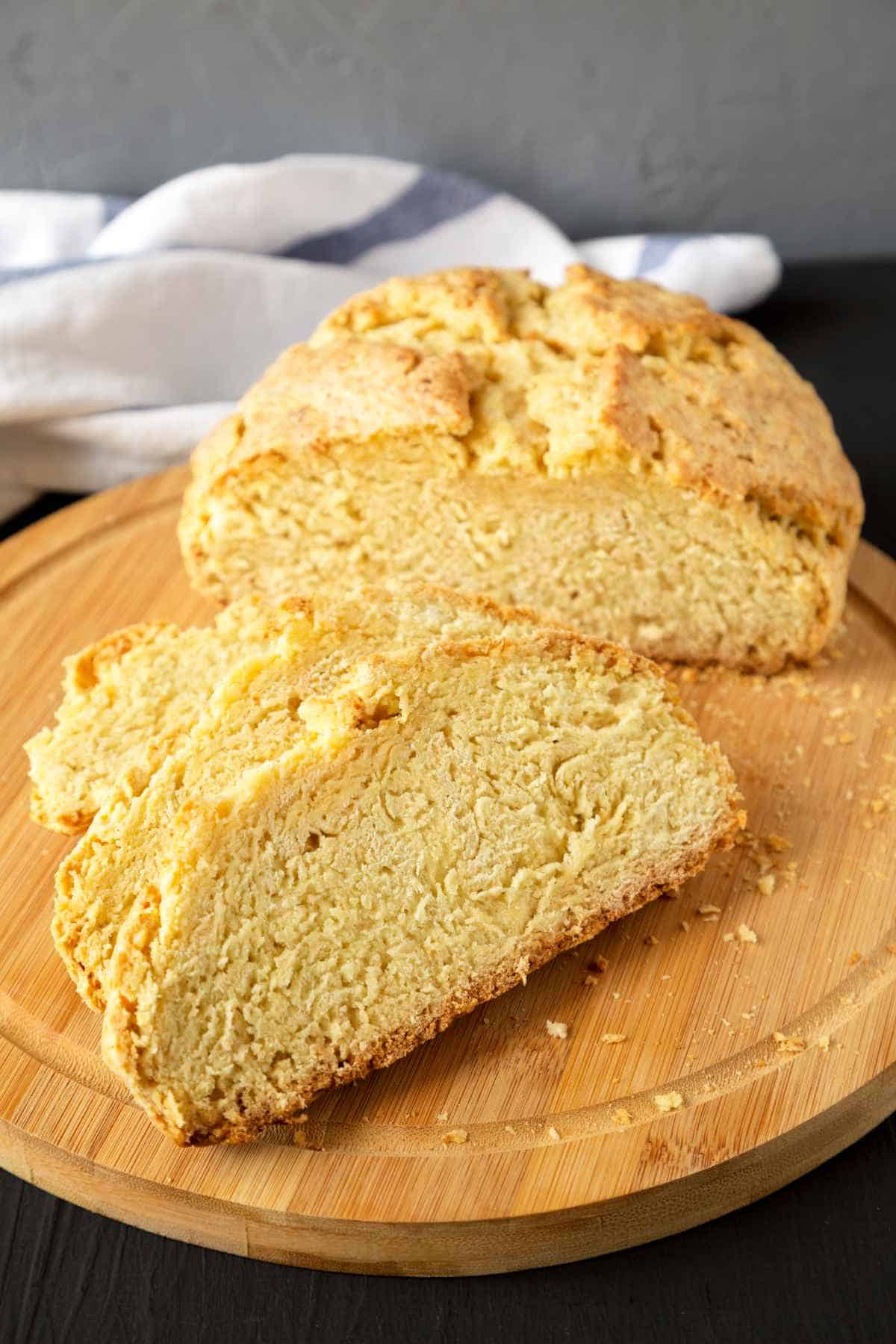 A round loaf of golden-brown gluten free Irish soda bread with a crunchy crust is sliced on a wooden cutting board, showing its dense, crumbly interior. A striped cloth is in the background.