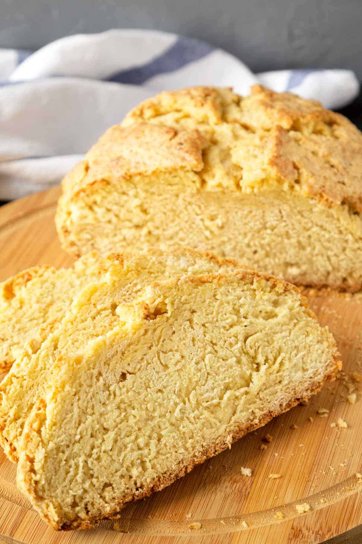 A round loaf of golden-brown gluten free Irish soda bread with a crunchy crust is sliced on a wooden cutting board, showing its dense, crumbly interior. A striped cloth is in the background.