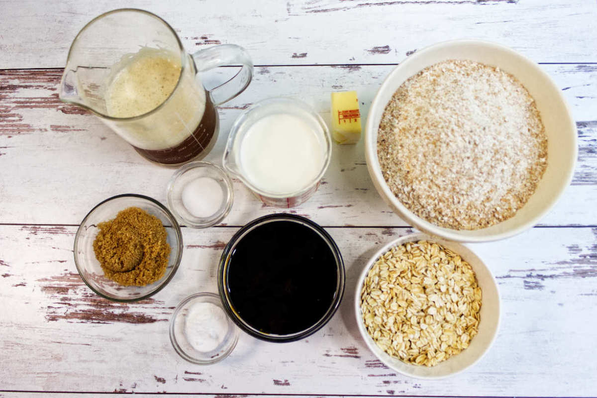 A top-down view of Irish Brown Bread ingredients in bowls and a measuring cup, including brown sugar, salt, baking soda, molasses, oats, wheat bran, milk, and butter, arranged on a white wooden surface.