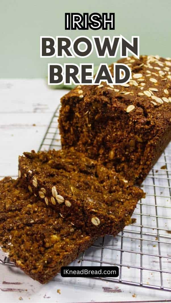 A loaf of Irish brown bread topped with oats sits on a cooling rack. Two thick slices are cut from the loaf, revealing a dense, textured crumb. Text reads "Irish Brown Bread" and "IKneadBread.com.