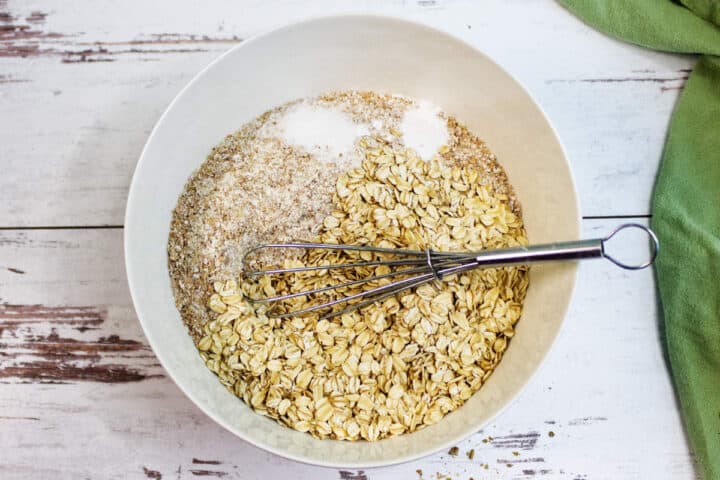 A white bowl contains oats, wheat bran, and salt-classic ingredients for Irish Brown Bread-with a metal whisk resting inside. The bowl sits on a rustic white wooden surface next to a green cloth.