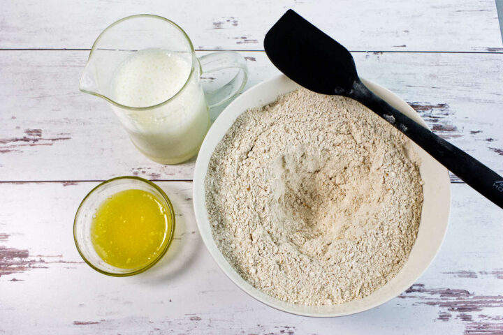 A bowl of flour with a black spatula, a glass measuring cup of milk, and a small bowl of melted butter on a white wooden surface—perfect essentials for making traditional Irish Brown Bread.