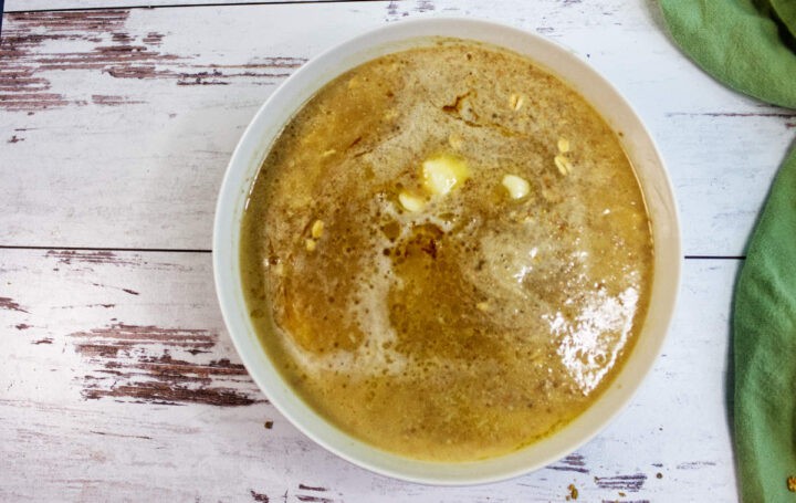 A bowl of hot soup or broth with melted butter on top, placed on a rustic white wooden surface next to a green cloth and a slice of Irish Brown Bread.
