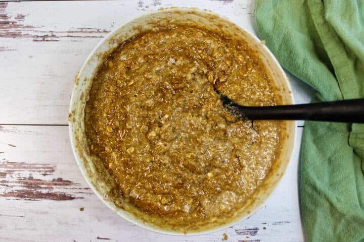 A mixing bowl filled with wet oat and Irish Brown Bread batter mixture, being stirred with a black spatula, sits on a white wooden surface next to a green cloth.