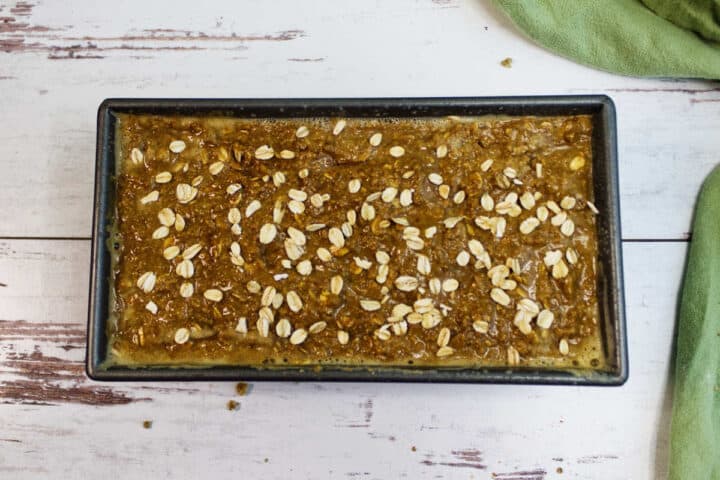 A rectangular baking pan filled with Irish Brown Bread mixture, golden-brown and topped with scattered rolled oats, sits on a white wooden surface next to a green cloth.