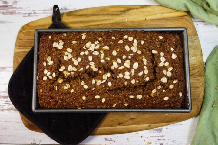 A rectangular loaf of Irish Brown Bread topped with oats sits in a metal pan on a wooden cutting board, with a black oven mitt and a green cloth nearby.