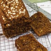 A loaf of Irish Brown Bread topped with rolled oats sits on a wire cooling rack, with two slices cut and lying next to it. A bread knife is partially visible nearby.