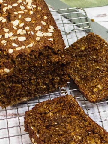 A loaf of Irish Brown Bread topped with rolled oats sits on a wire cooling rack, with two slices cut and lying next to it. A bread knife is partially visible nearby.