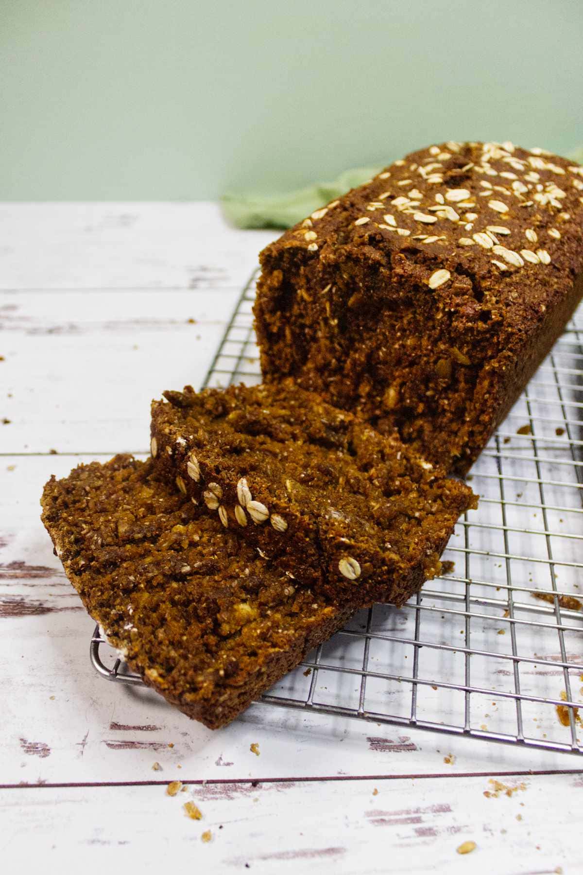 A loaf of Irish Brown Bread with oats sprinkled on top sits on a cooling rack. Two slices have been cut, revealing a moist texture inside. The bread rests on a white wooden surface with some crumbs scattered around.