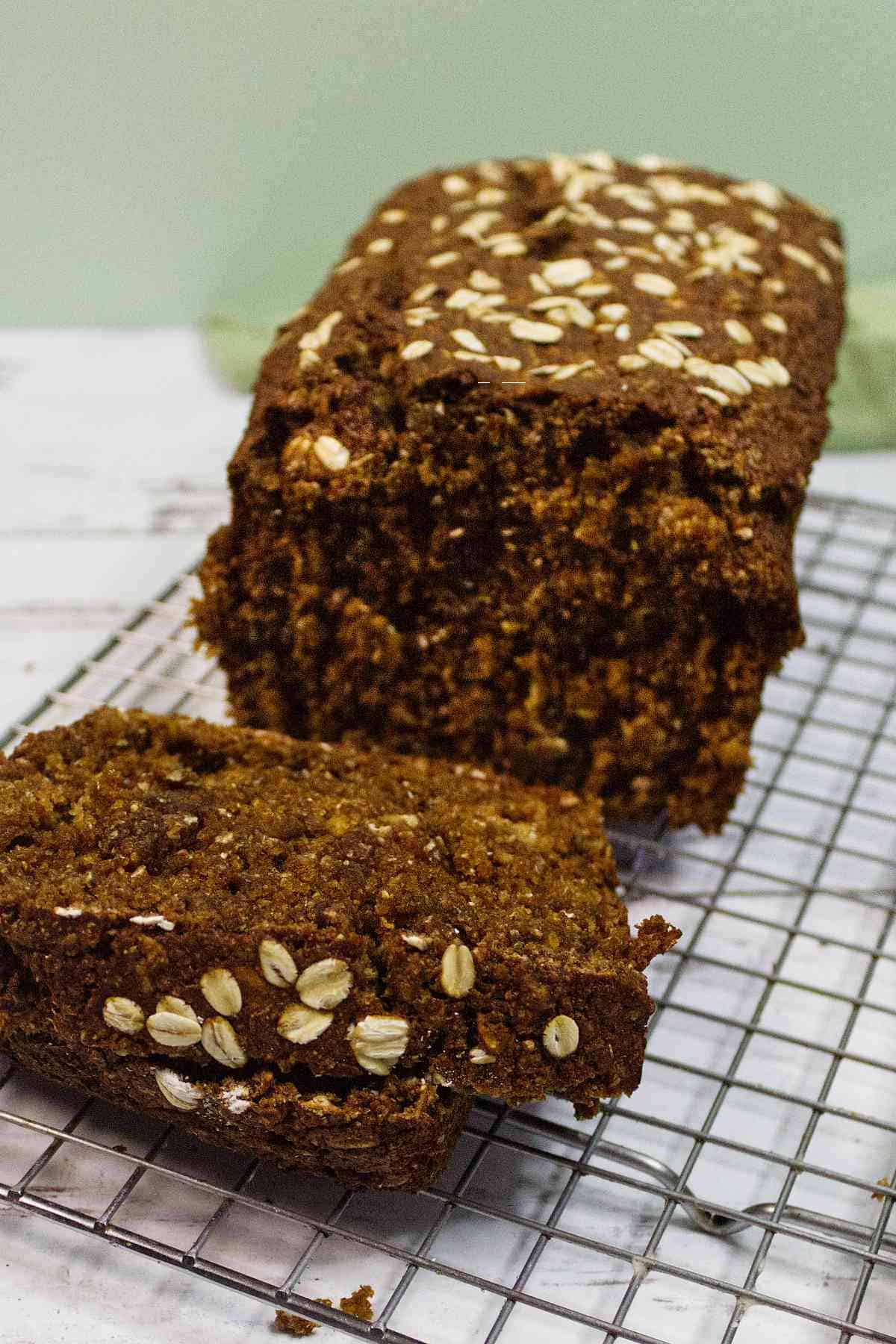 A loaf of Irish Brown Bread sits on a cooling rack, with a single slice cut and lying in front. The bread&rsquo;s top is sprinkled with whole oats. The background is a light surface with a pale green wall.