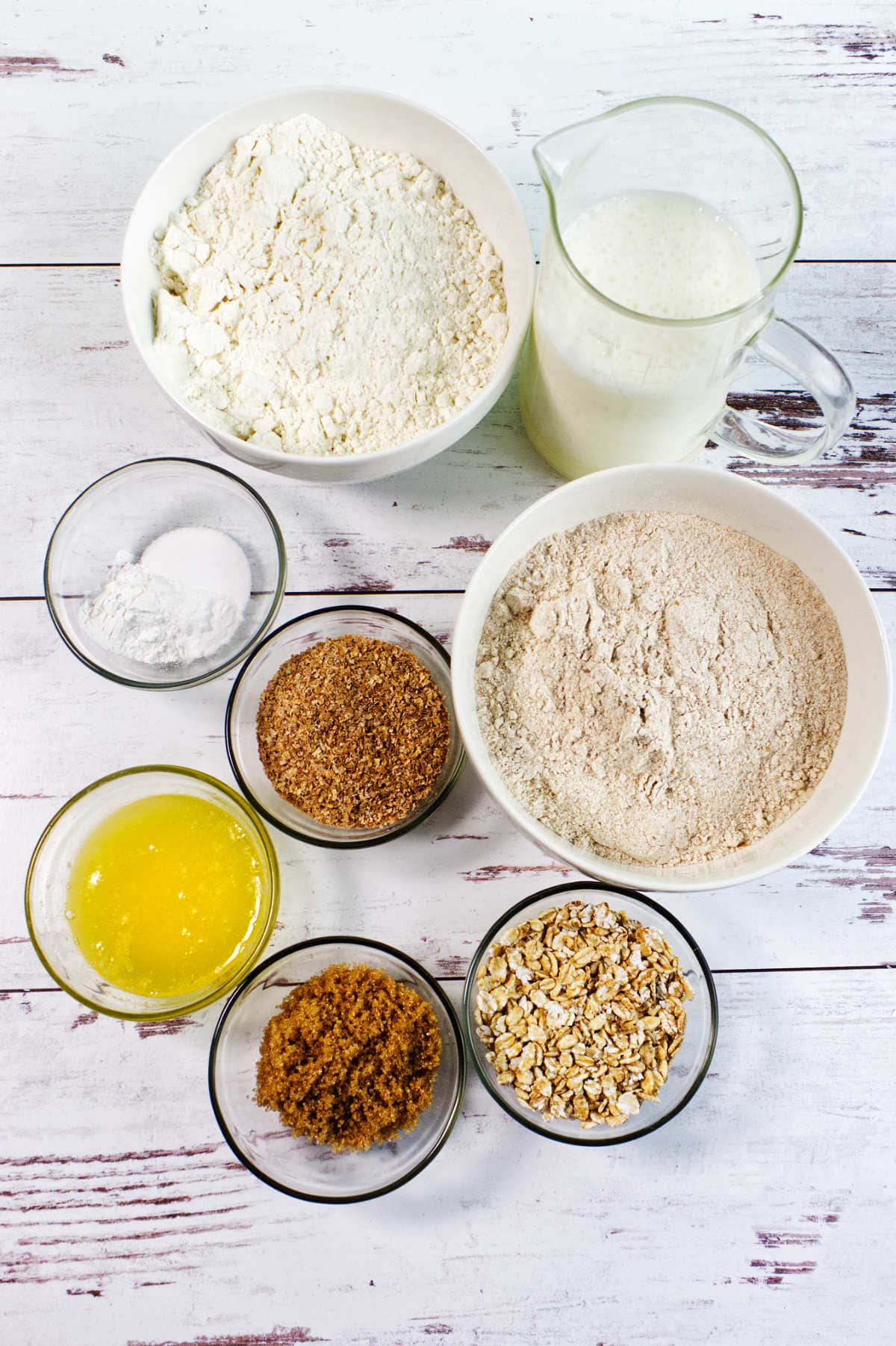 Eight bowls and a pitcher on a white surface, each filled with key irish wheaten bread ingredients: flour, milk, baking soda, melted butter, ground flaxseed, brown sugar, oats, and whole wheat flour.