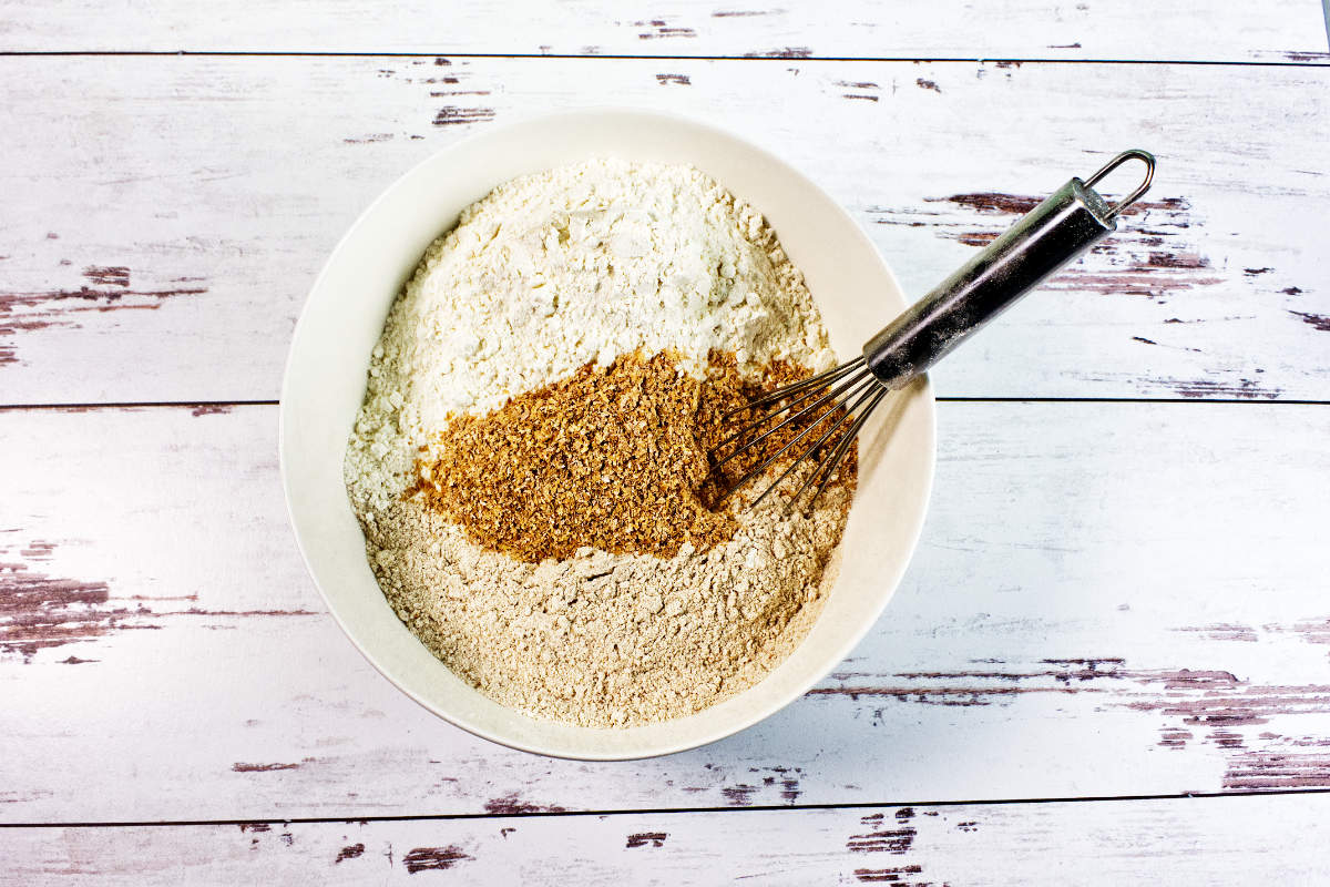 A white bowl filled with flour and brown sugar being mixed with a metal whisk, ready for making Irish wheaten bread, sits on a rustic white wooden surface.