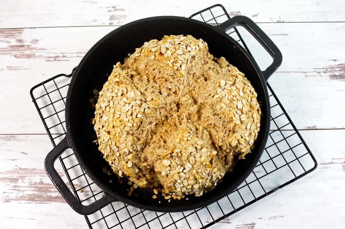A round loaf of homemade whole grain bread topped with oats, resting in a black cast iron pot on a cooling rack over a white wooden surface.