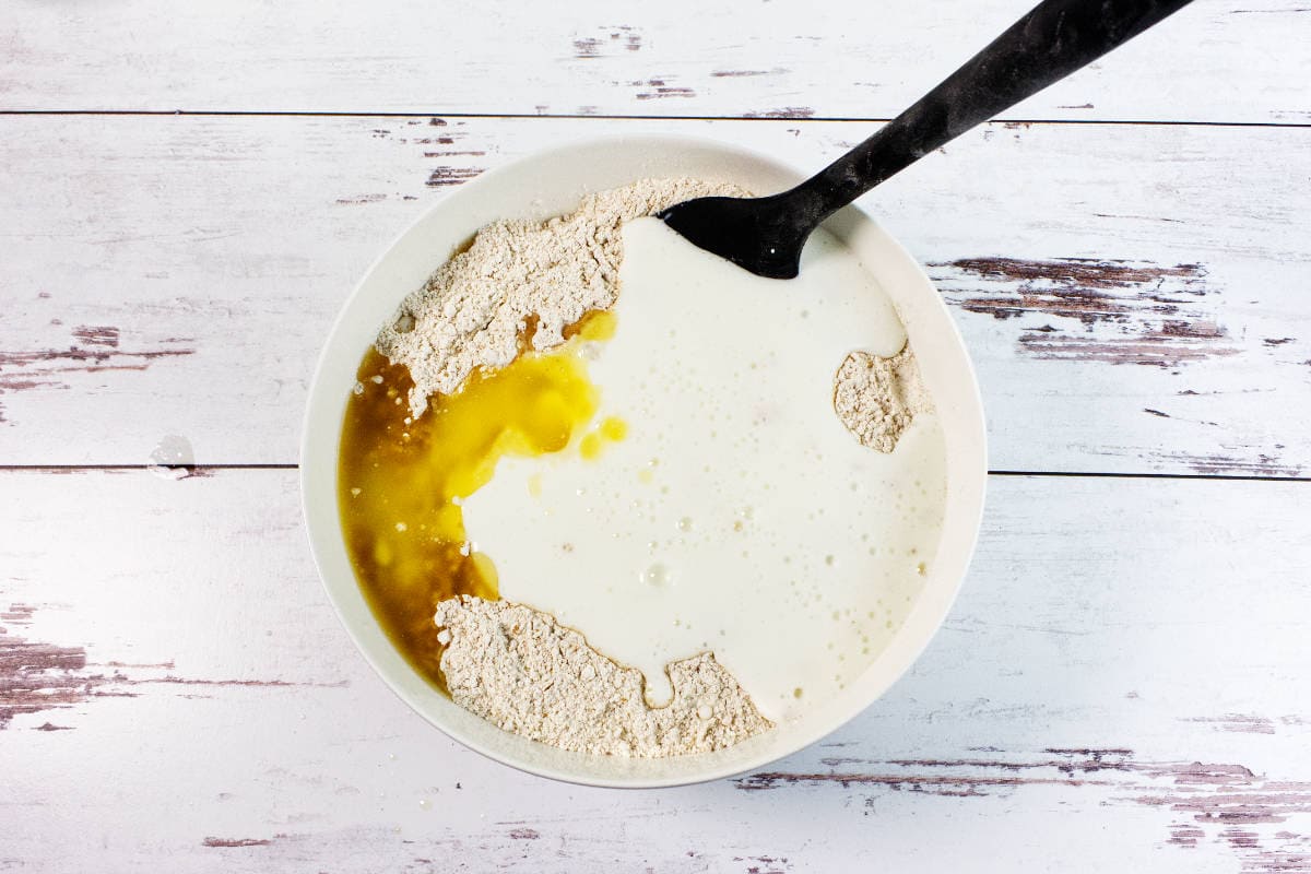 A white bowl containing ingredients for Irish wheaten bread-flour, eggs, milk, and vanilla-being mixed with a black spoon, sitting on a white wooden surface.