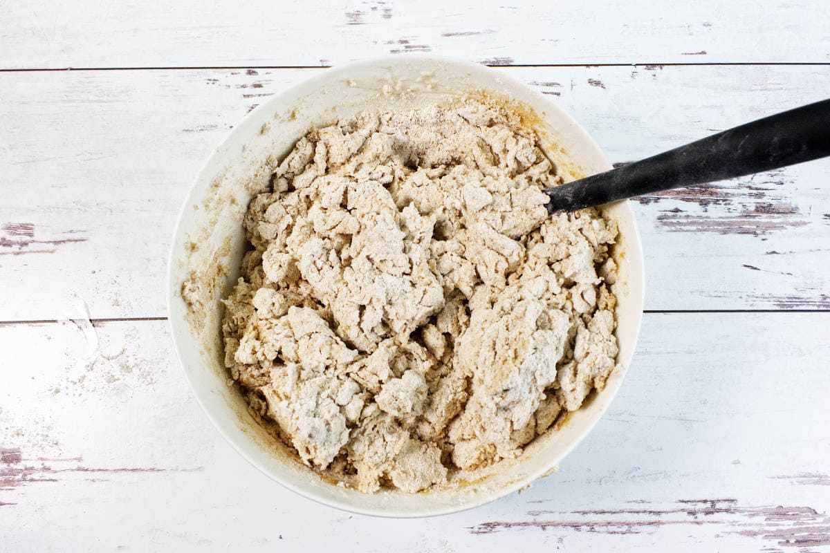 A large white bowl filled with crumbly, partially mixed Irish wheaten bread dough sits on a white wooden surface. A black mixing spoon rests inside the bowl.