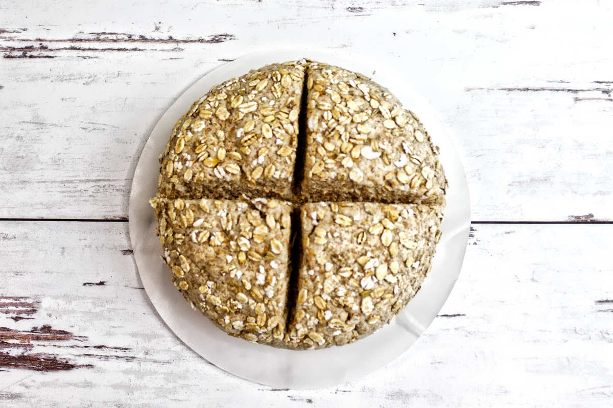 A round loaf of Irish wheaten bread, oat-topped and sliced into four equal pieces, sits on a white plate against a rustic white wooden background.