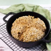A round loaf of rustic Irish wheaten bread sits in a black cast iron pot on a cooling rack. The bread is topped with rolled oats and has a rough, golden-brown crust. A green cloth rests in the background.
