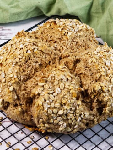 A round loaf of Irish wheaten bread, topped with oats, sits on a black cooling rack, with a green cloth and white table surface in the background.
