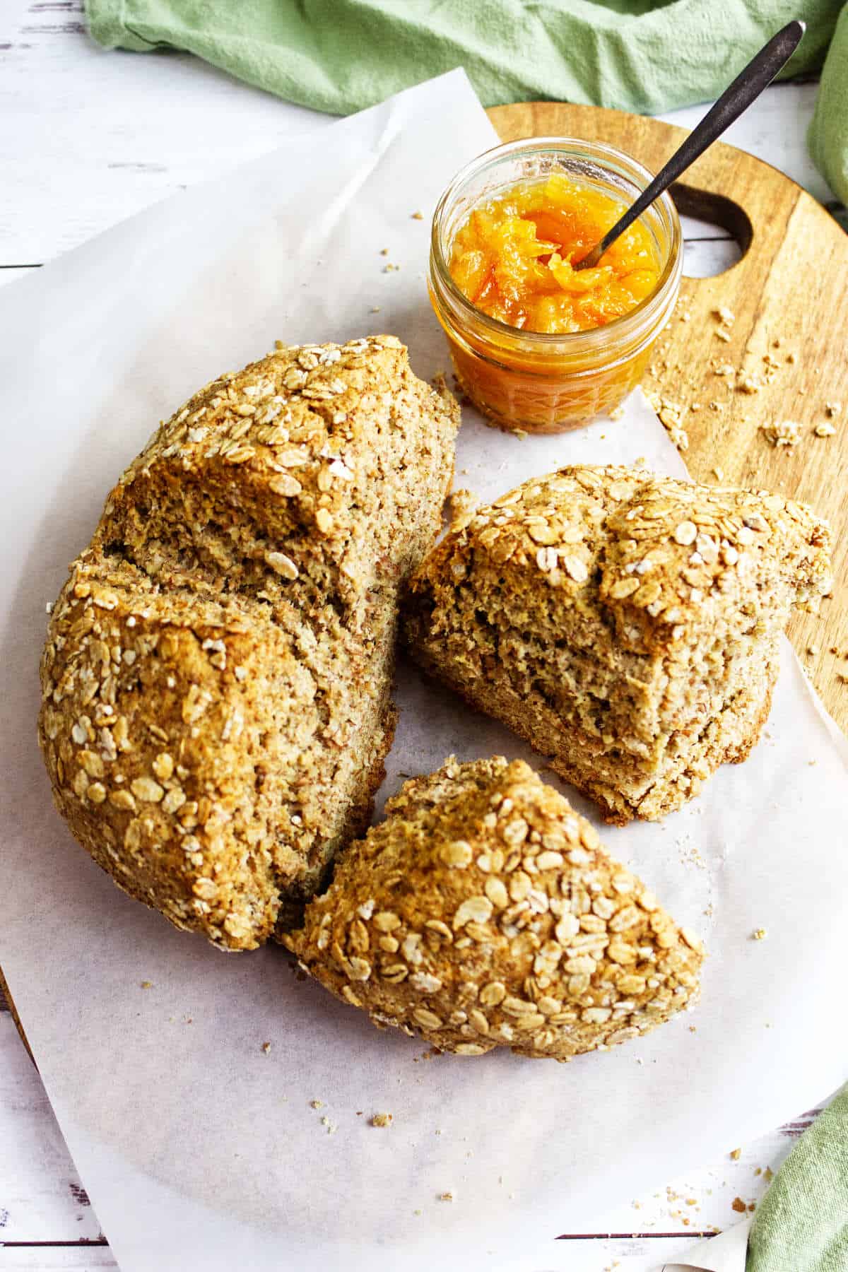 A loaf of oat-topped Irish wheaten bread is sliced into quarters on parchment paper, with a jar of orange marmalade and a spoon nearby, all set on a wooden board.