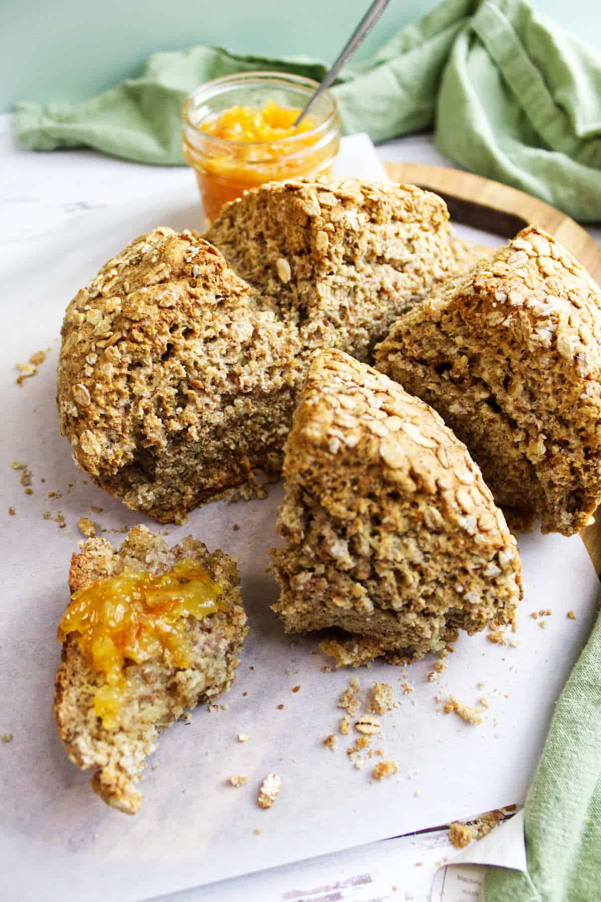A sliced round loaf of rustic Irish wheaten bread sits on parchment paper, with one piece spread with orange marmalade. A jar of marmalade and a green cloth are in the background, and crumbs are scattered around.