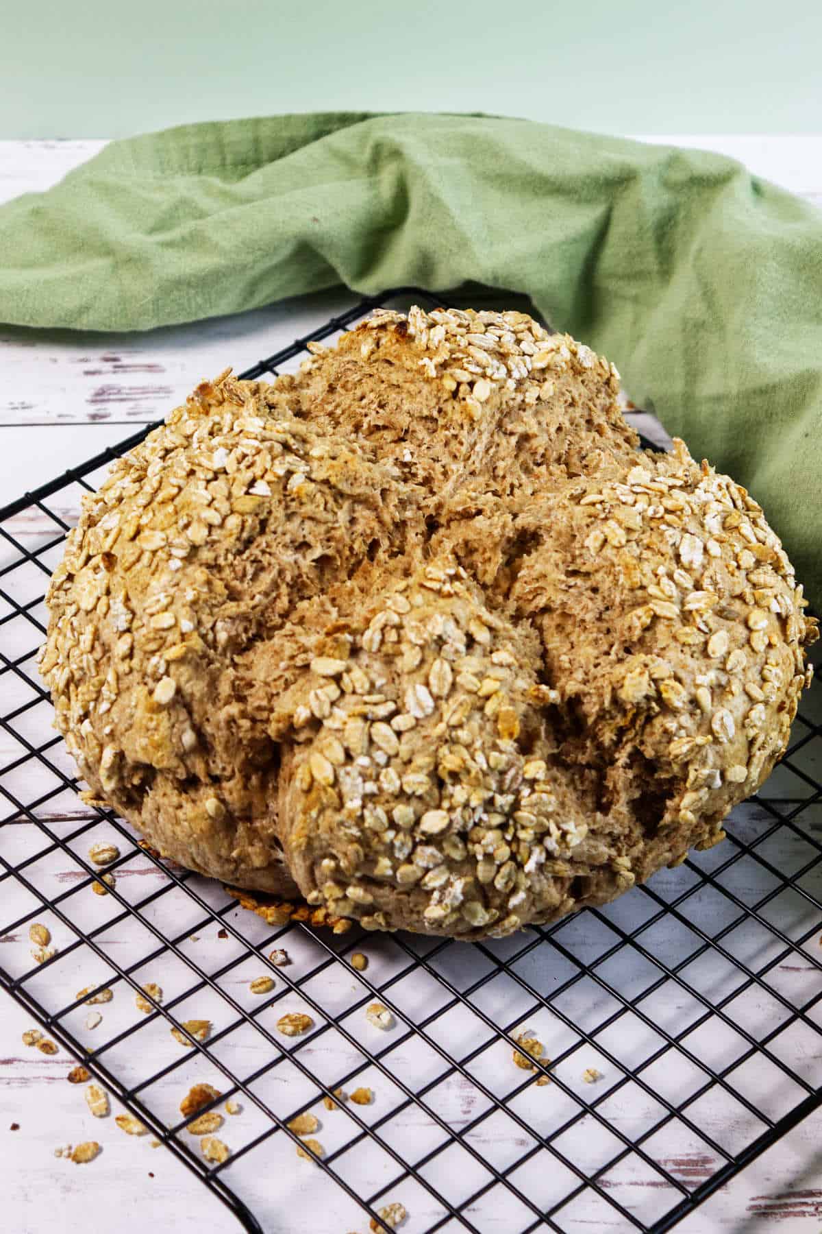 A round loaf of Irish wheaten bread topped with oats sits on a cooling rack, scattered oats around it. A green cloth is in the background on a white wooden surface.