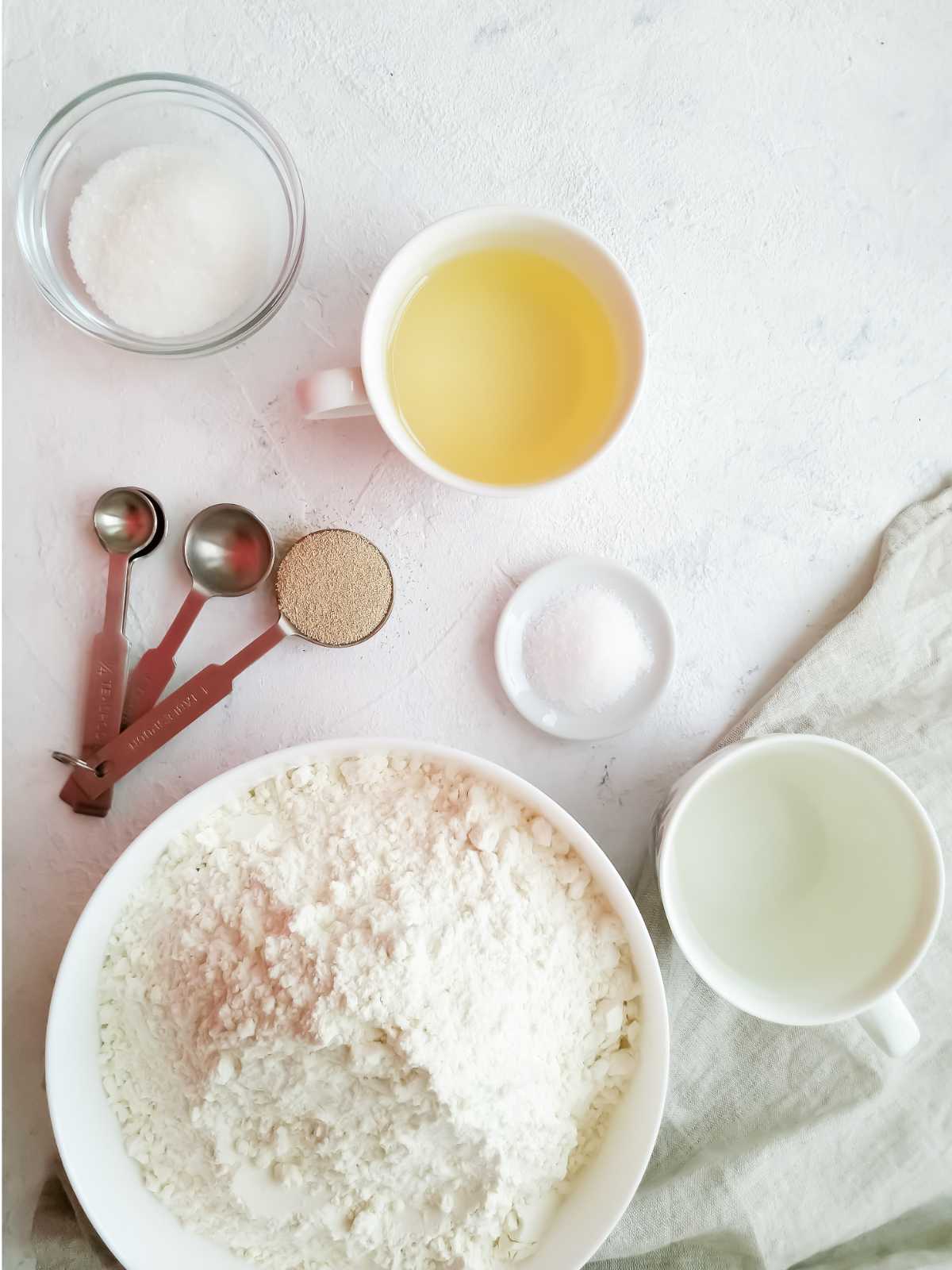 Ingredients for mantou buns are arranged on a light surface: flour in a white bowl, a cup of yellow liquid, clear liquid, sugar in a glass bowl, salt, measuring spoons, and yeast-all set beside a napkin.