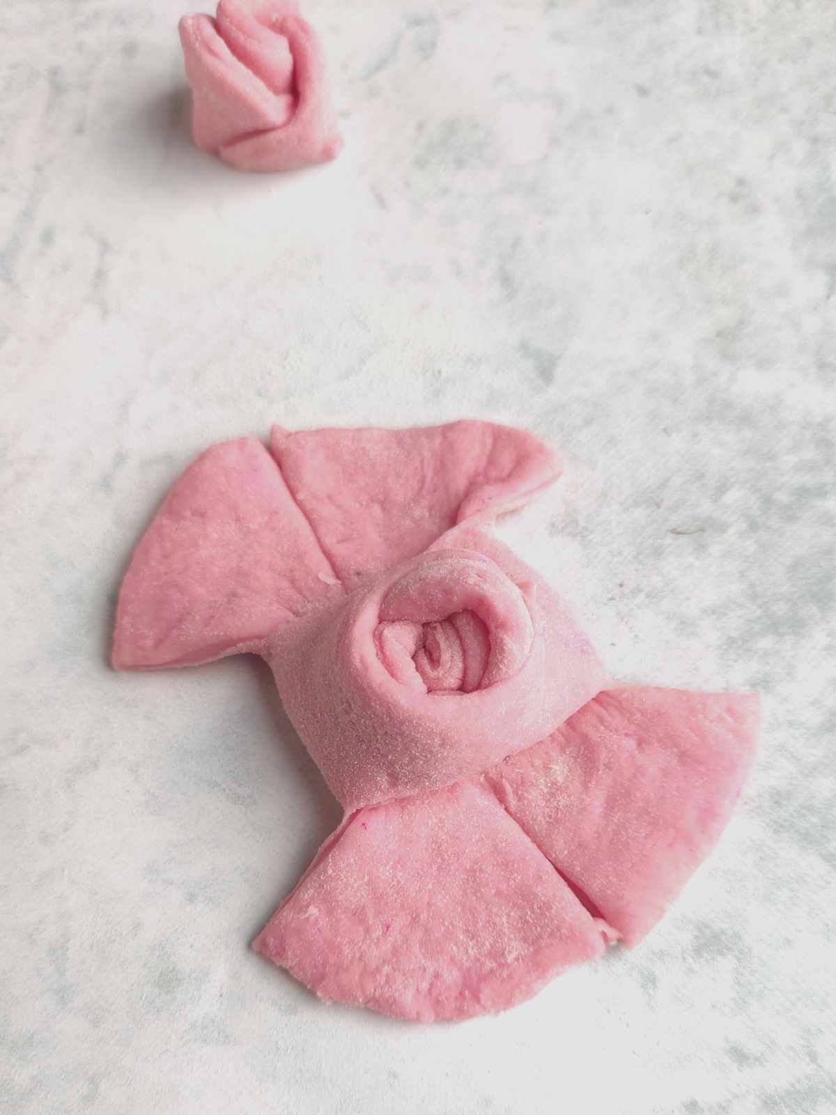 A close-up of pink mantou buns shaped like roses, with petal-like cuts. One finished rose and one partially formed bun rest on a light, textured surface.