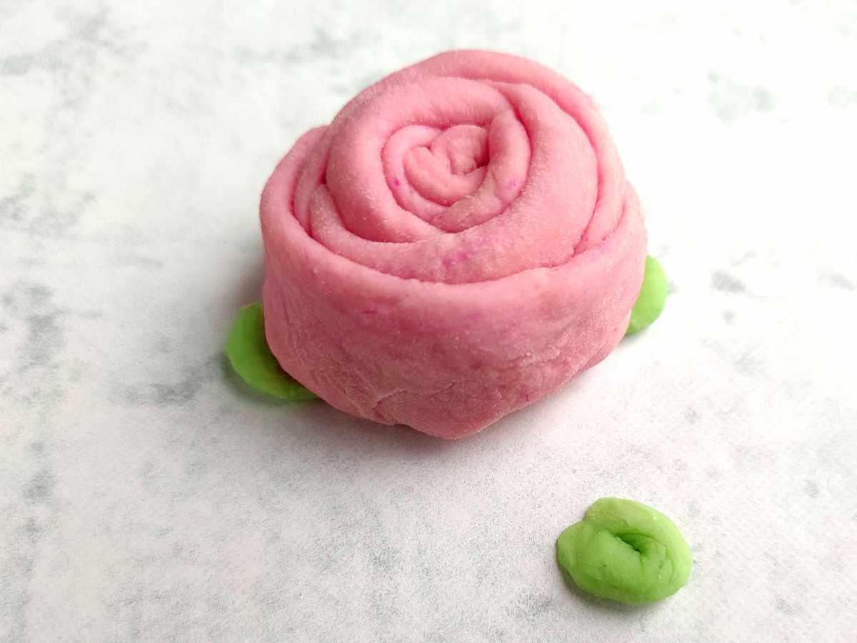 A pink rose-shaped mantou bun with two small green leaf decorations sits on a light, textured surface.