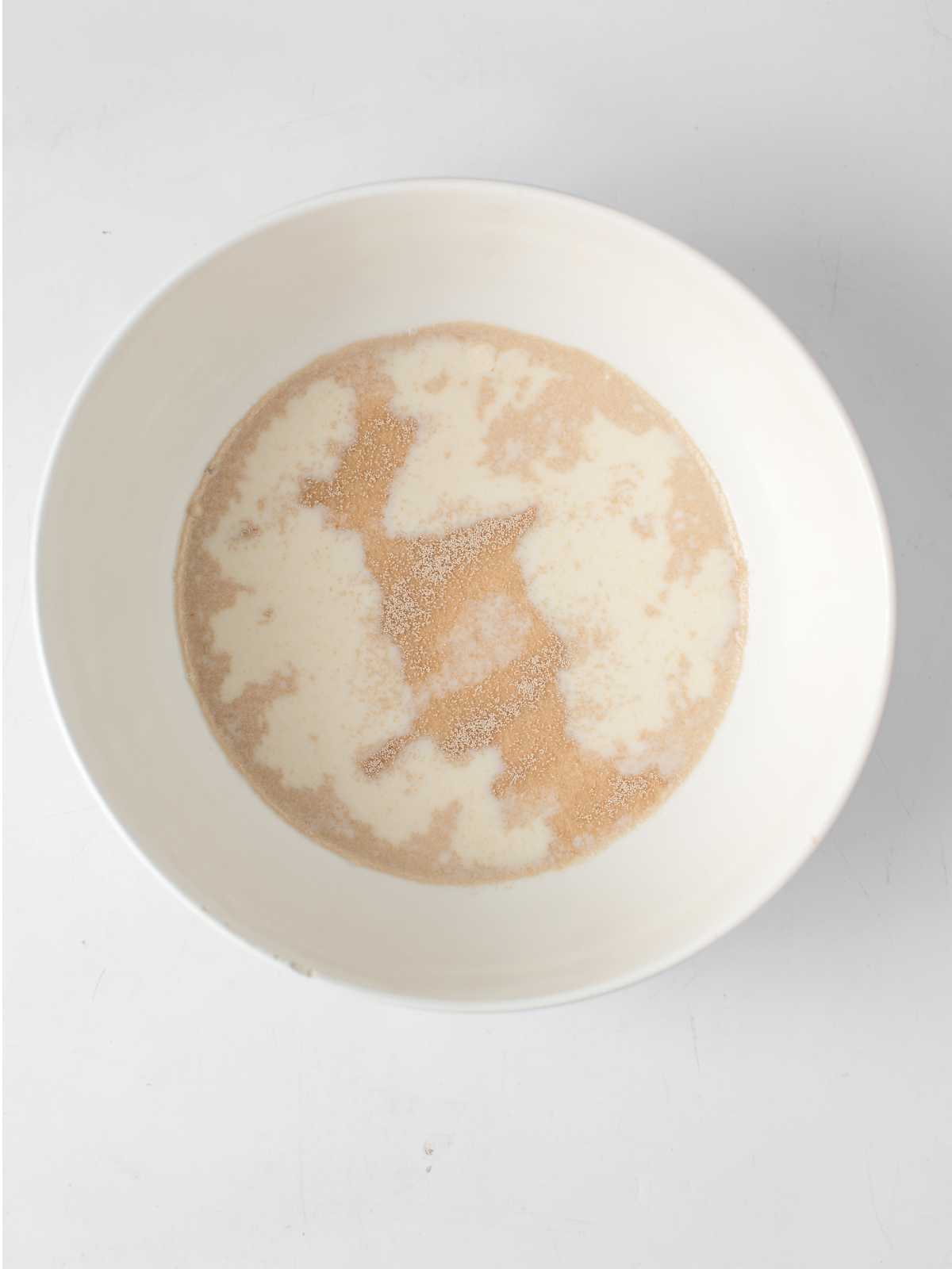 A white bowl containing a mixture of milk and yeast granules, the first step in preparing fluffy mantou buns, rests on a white surface.