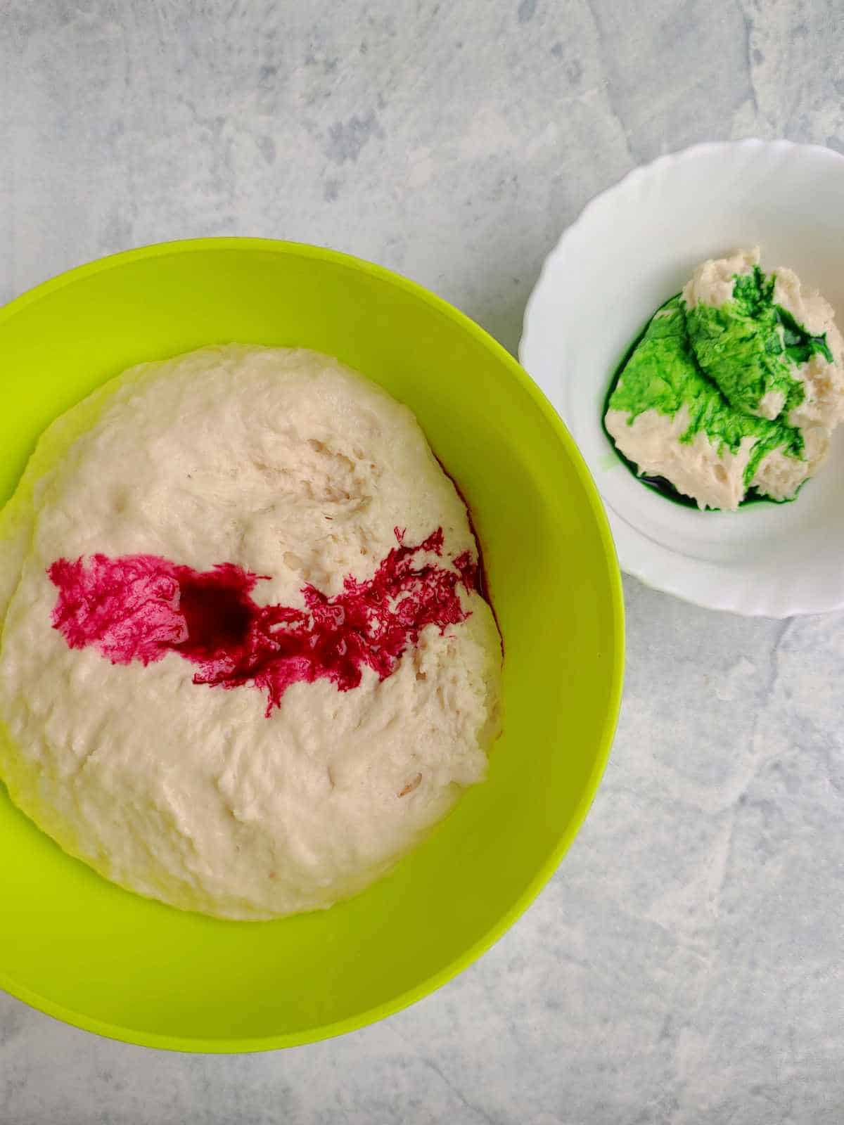A yellow bowl with dough and a streak of red food coloring, next to a white plate holding a piece of mantou bun dough partially covered in green food coloring, on a light surface.