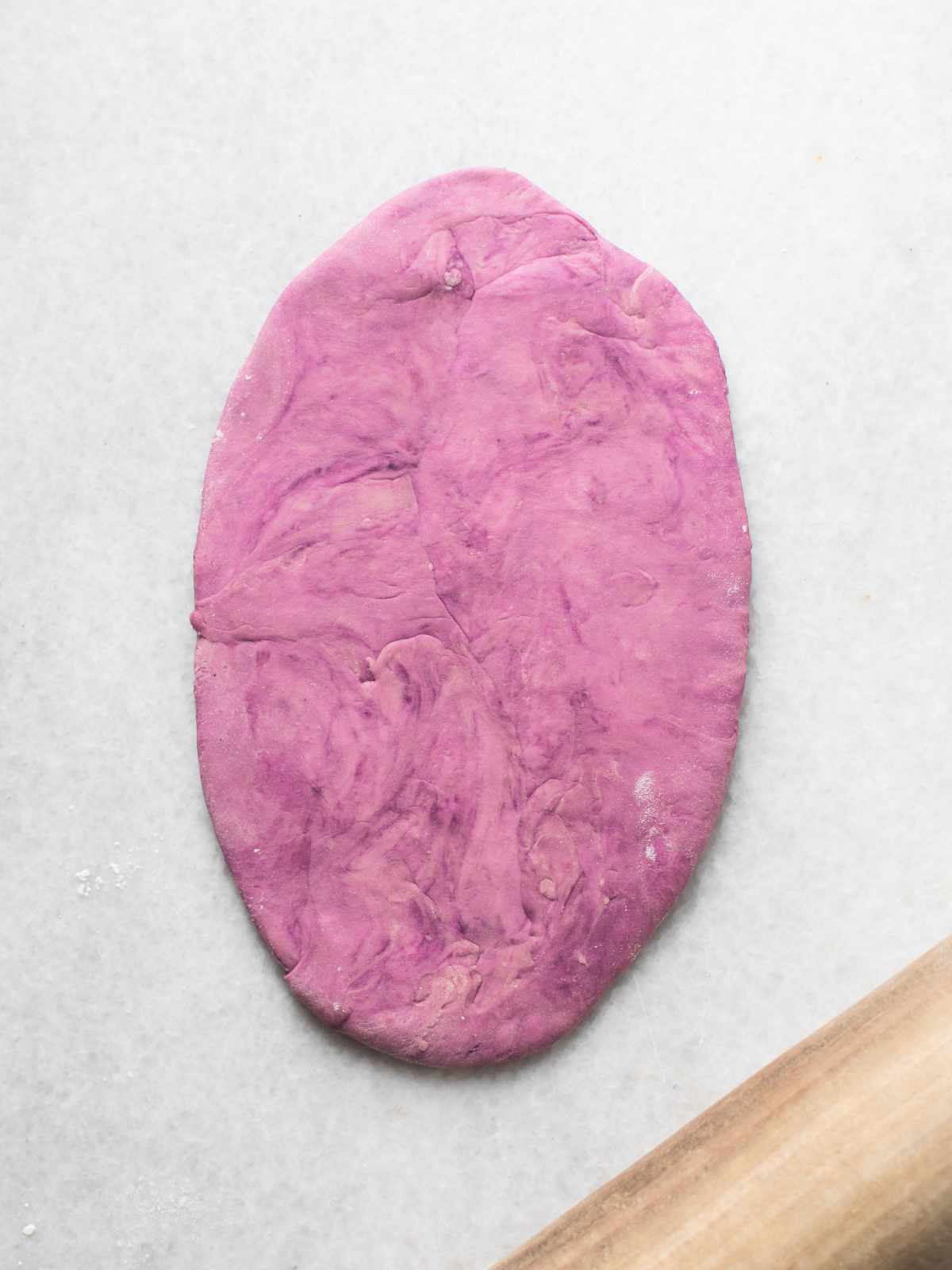 An oval-shaped piece of purple dough, soon to become mantou buns, is rolled out on a light surface, with the edge of a wooden rolling pin visible in the bottom right corner.