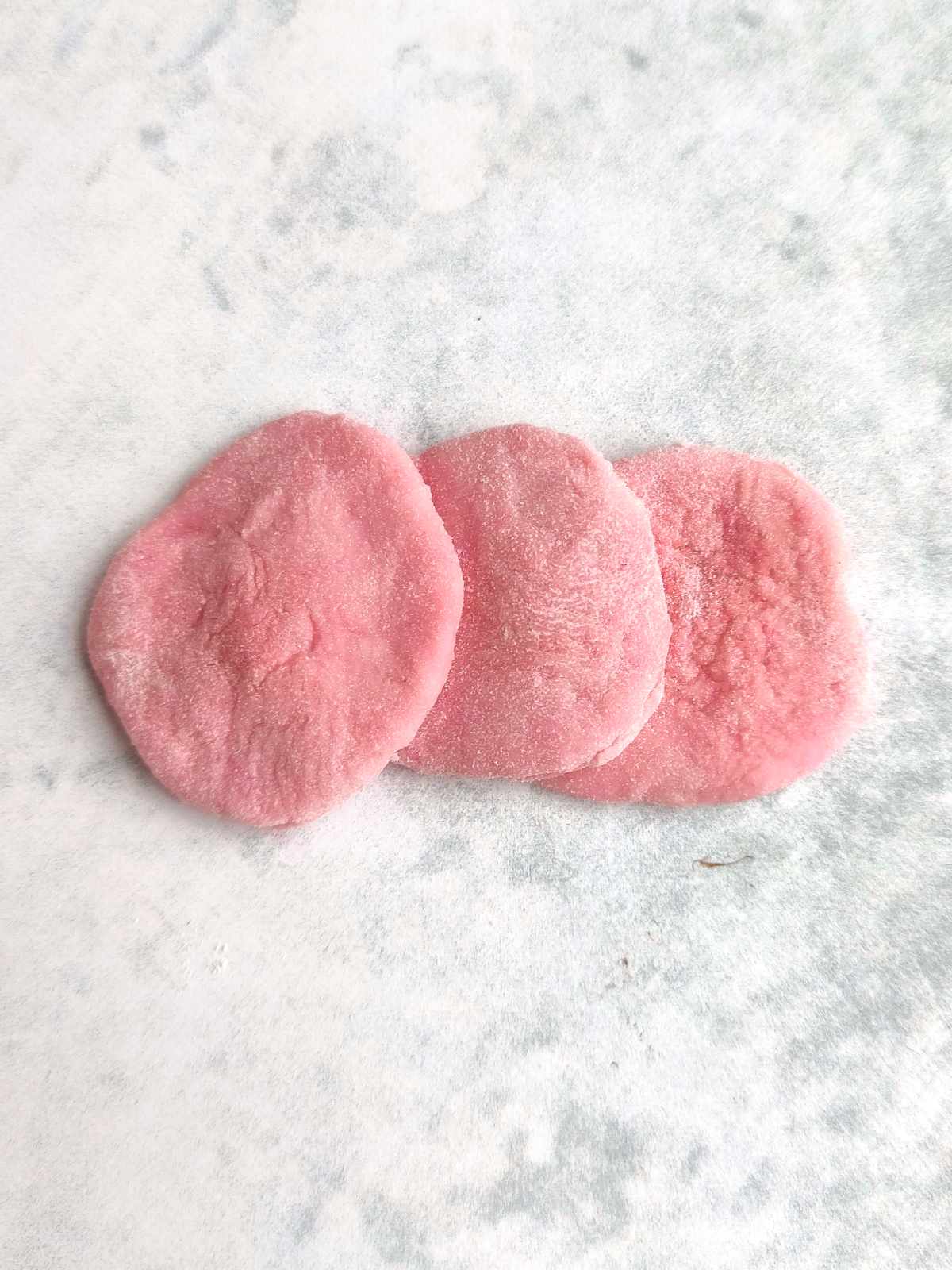 Three round, pink pieces of mochi lie side by side on a light, textured surface. Reminiscent of mantou buns, the mochi look soft and slightly powdery.